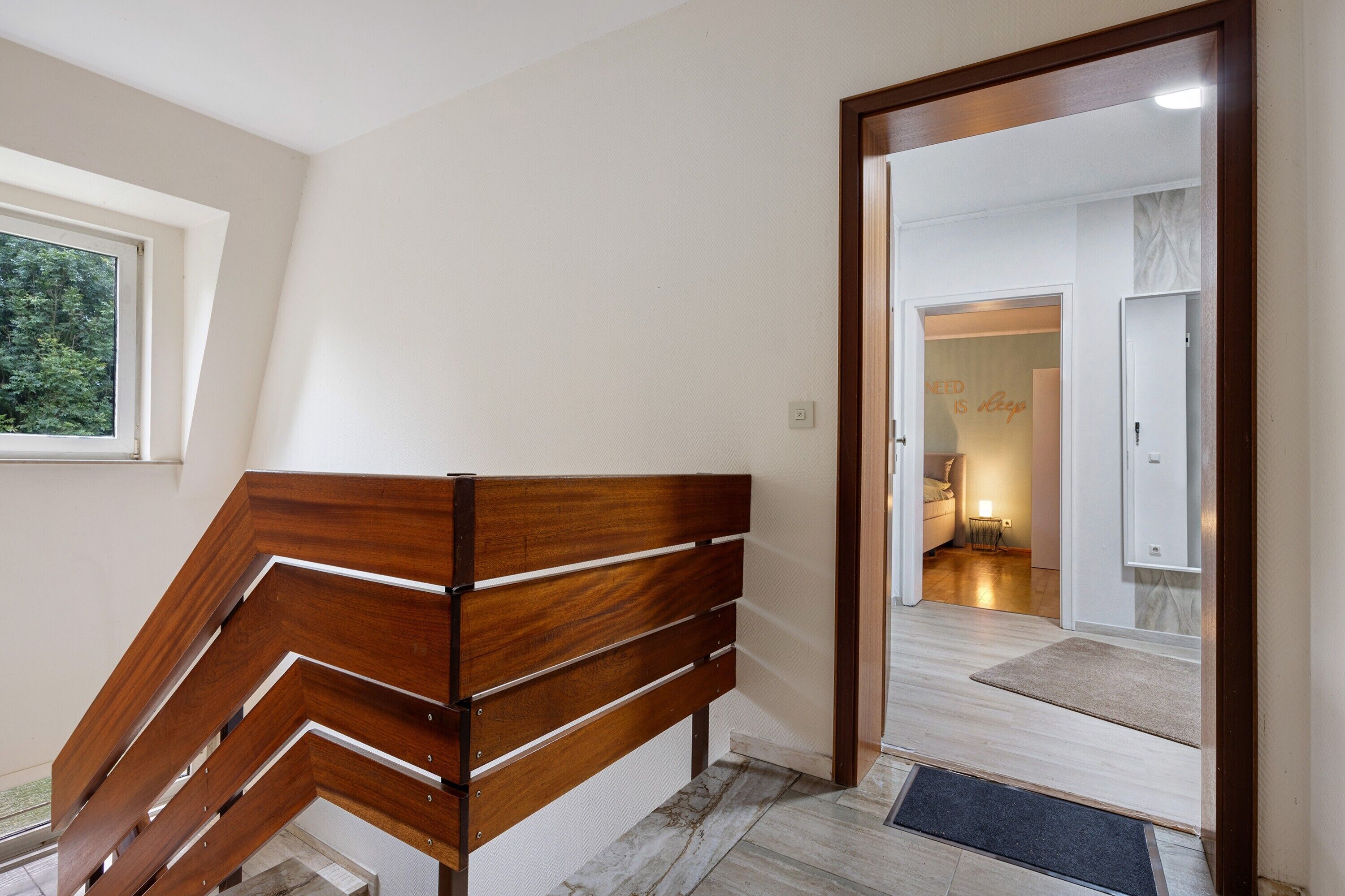Un escalier en bois au design moderne, menant à une pièce dont la porte est visible et où se trouve un petit tapis.