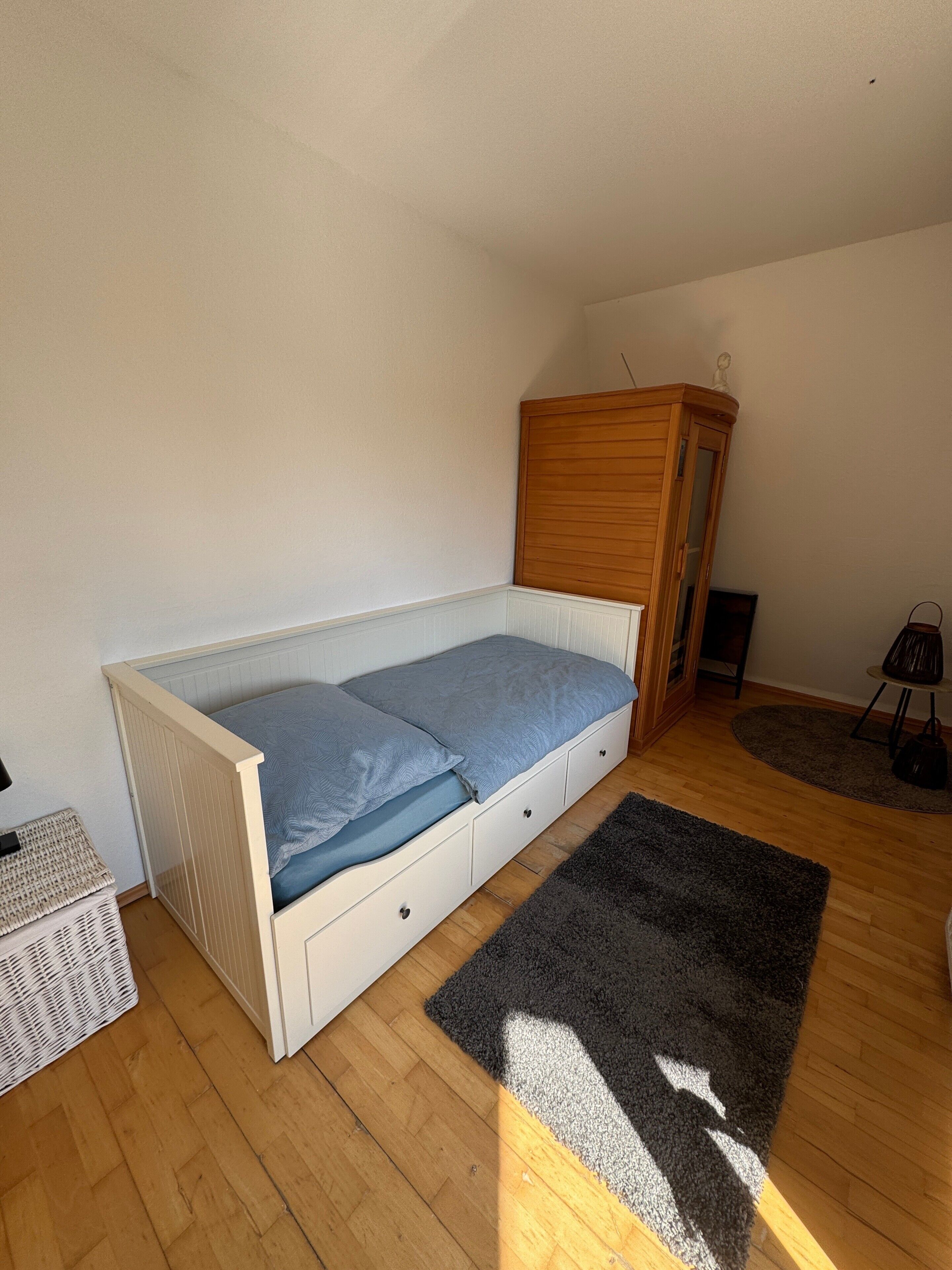 Una habitación pequeña con cama individual, un armario de madera, un radiador blanco y una alfombra gris sobre piso de madera.