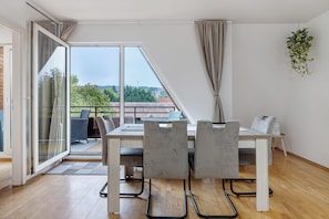 Un área de comedor con una mesa y sillas, un puertas corredera de vidrio que da a un balcón exterior, y una planta colgante.