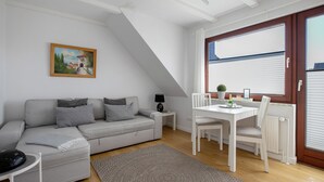 A modern living room with a grey sofa, a dining table with chairs, a small side table with a lamp, and a painting on the wall. - Apartment 'Edda 3' with Shared Garden, Balcony and Wi-Fi (Meldorf)