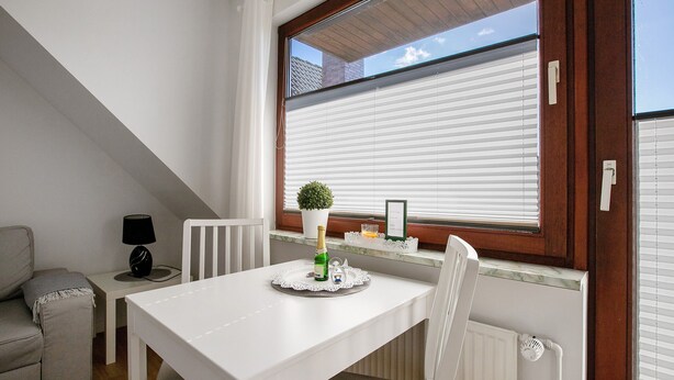 A white dining table with a bottle, glass, and a small plant on a tray, a white chair, a white sofa, a small side table with a lamp, and a window with blinds. - Apartment 'Edda 3' with Shared Garden, Balcony and Wi-Fi (Meldorf)