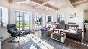 A spacious living room with a large sofa, a leather chair, and a coffee table. There are large windows with a view of the outdoors. - Holiday Home 'Lüthjes Friesenhaus' with Private Terrace, Garden and Wi-Fi (Rodenäs)