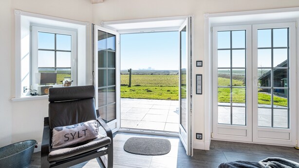 A room with a view of a green field through a glass door, a chair with a pillow, and a metal bucket. - Holiday Home 'Lüthjes Friesenhaus' with Private Terrace, Garden and Wi-Fi (Rodenäs)
