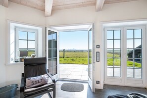 A room with a view of a green field through a glass door, a chair with a pillow, and a metal bucket. - Holiday Home 'Lüthjes Friesenhaus' with Private Terrace, Garden and Wi-Fi (Rodenäs)