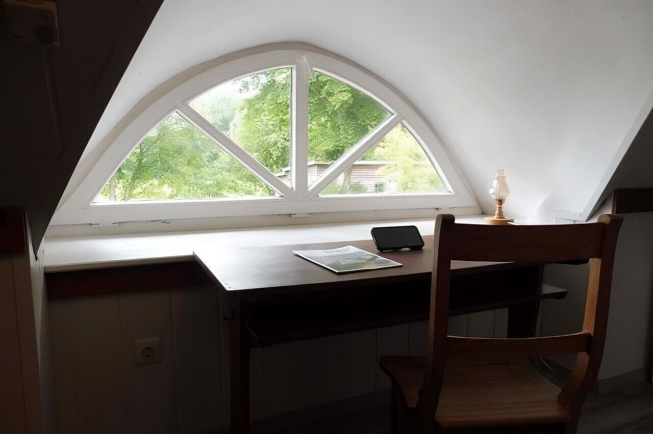Ein Zimmer mit schrägem Dach, einem Schreibtisch mit Stuhl, einem Fenster mit Blick auf Grünflächen und einer Lampe auf dem Schreibtisch.
