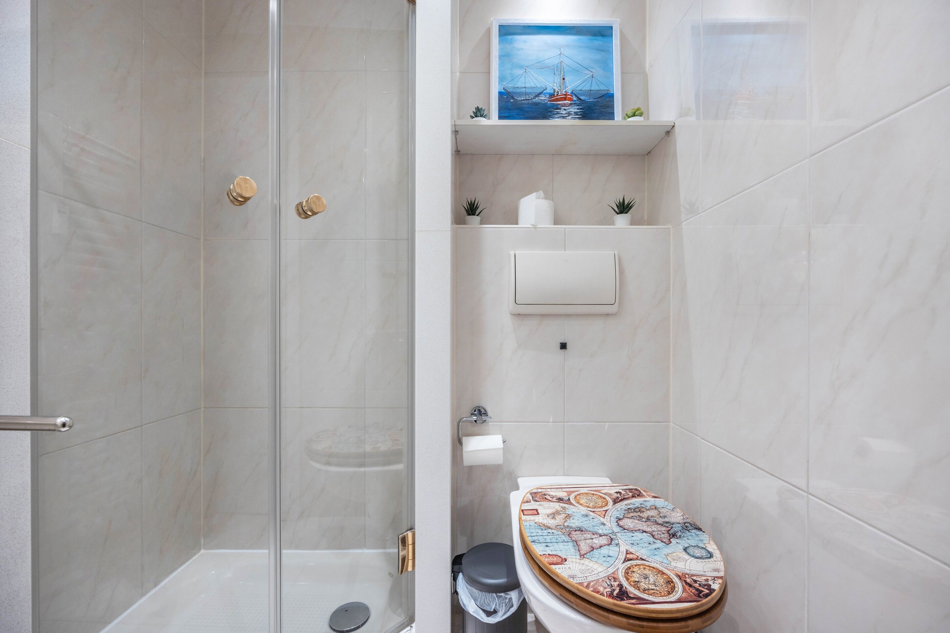 Une salle de bain avec une douche à parois de verre, des toilettes avec un abattant à motifs et une étagère ornée d’un tableau et de petites plantes.