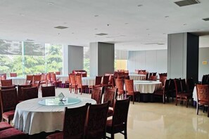 A spacious dining area with round tables and chairs, large windows, and a view of greenery.