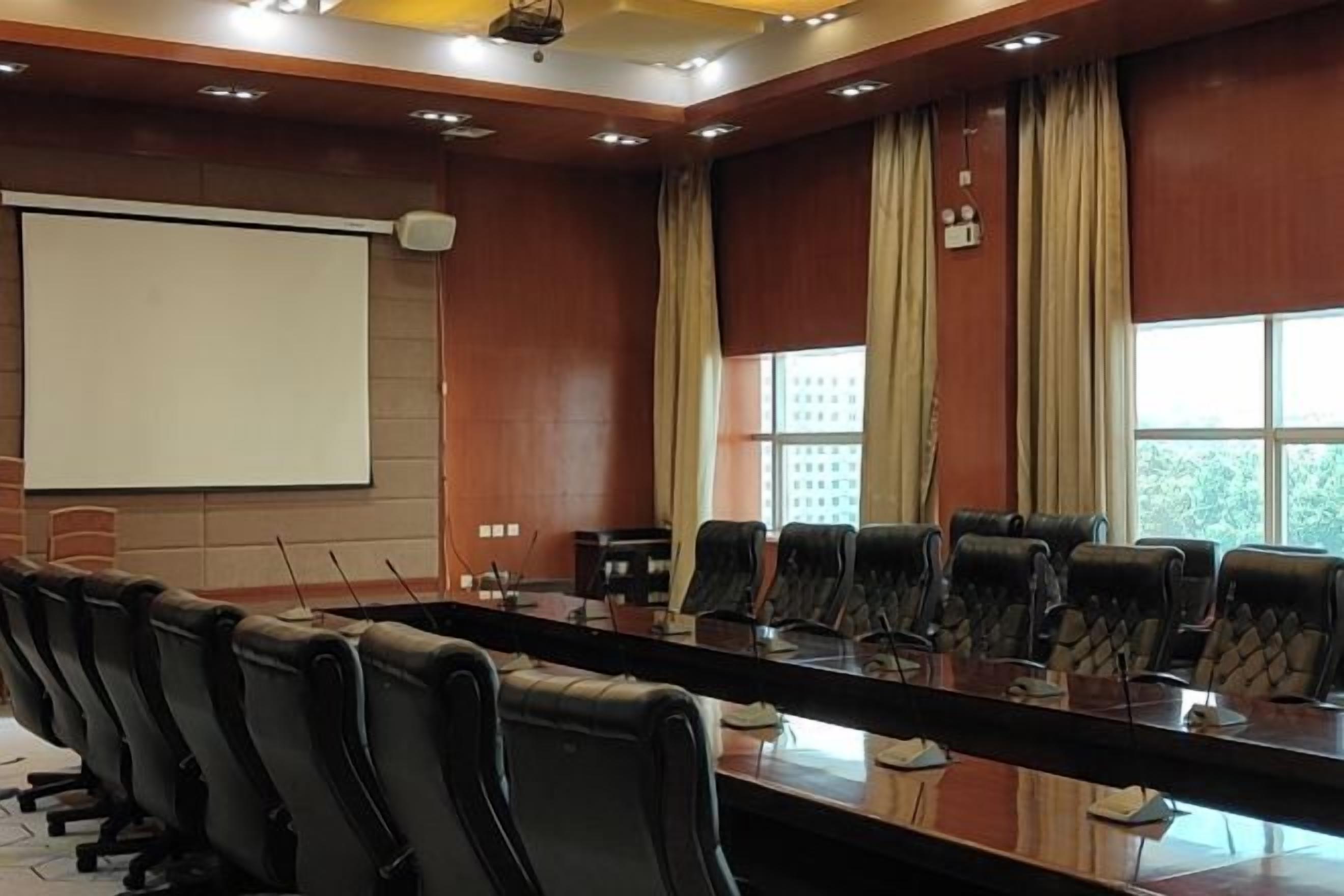 A conference room with a long table, multiple chairs, a projector screen, and large windows with curtains.