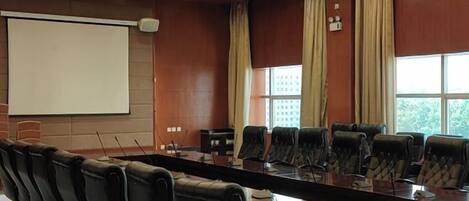 A conference room with a long table, multiple chairs, a projector screen, and large windows with curtains.