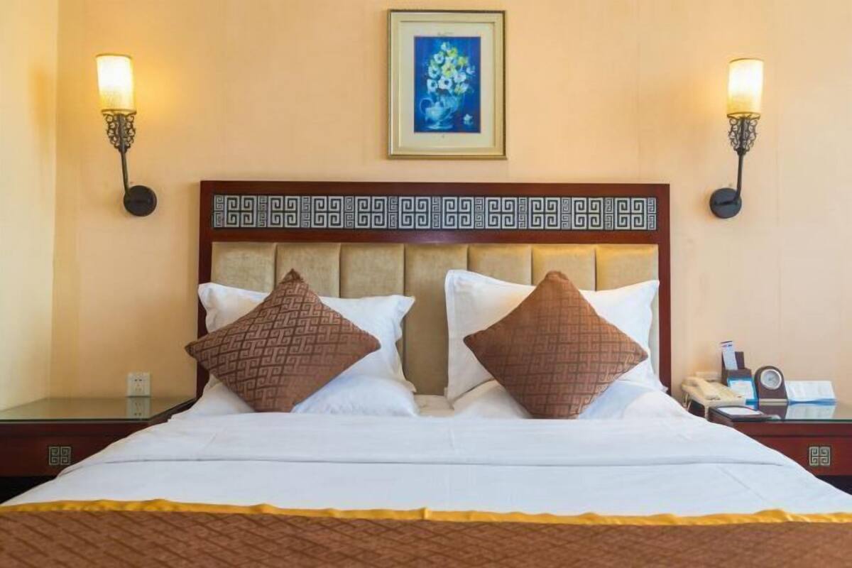 A hotel room with a large bed, two wall-mounted lamps, a framed artwork above the bed, and a bedside table with a telephone and a book.