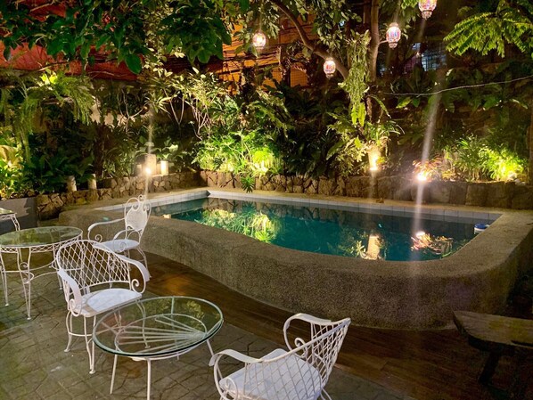 Zona de piscina con borde de piedra, rodeada de abundante vegetación y sillas de metal blanco.