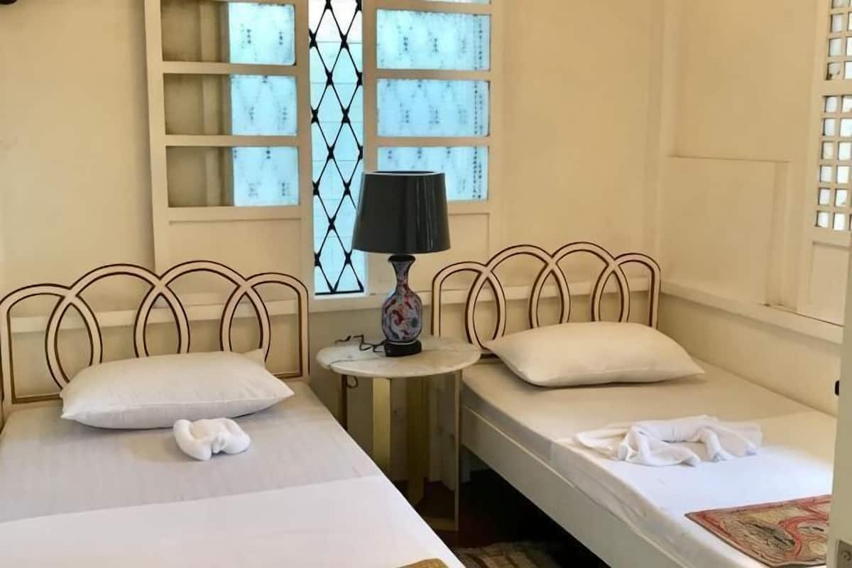 A room with two beds, a small table with a lamp, and a window with a decorative pattern.