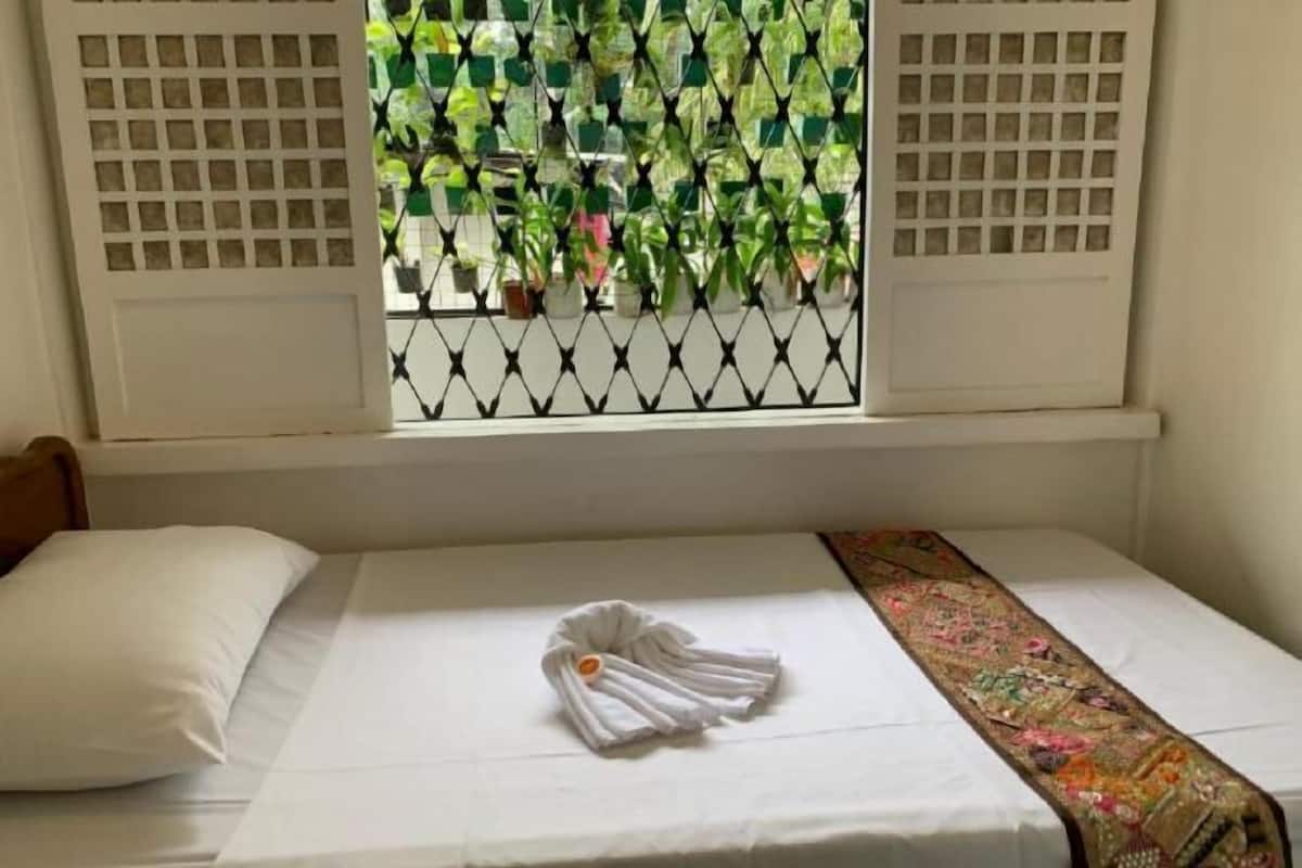 A bed with a white pillow and a folded towel, a patterned bedspread, and a window with a view of greenery outside.