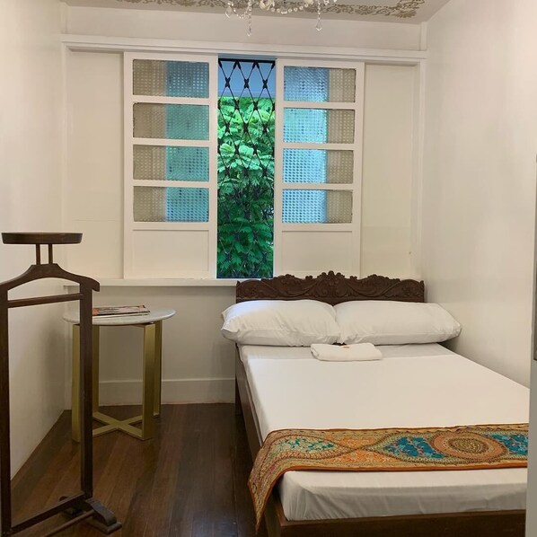 A bedroom with a bed, a small table, and a window with a view of green foliage. - Courtyard Hotel (Pasay)