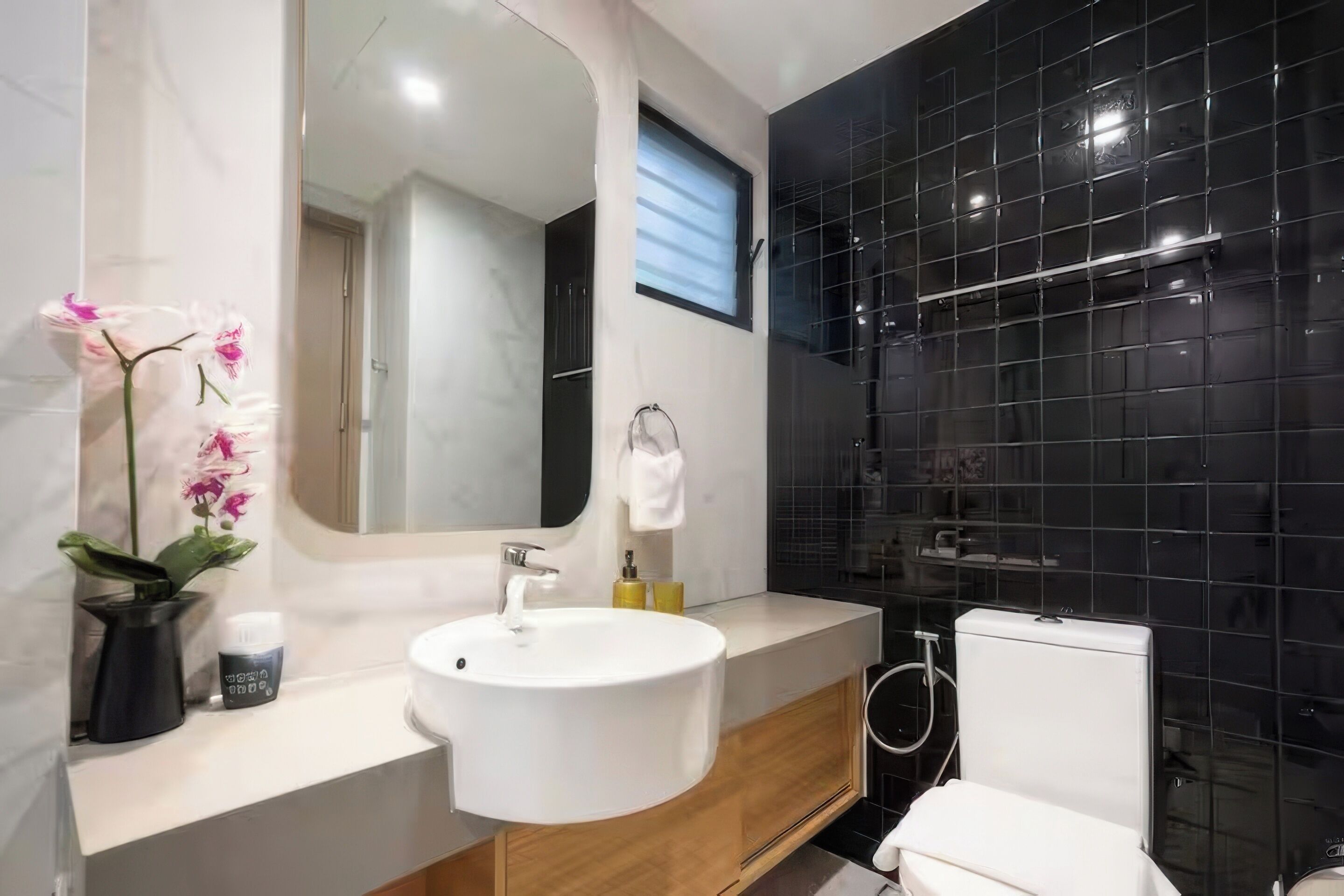 A modern bathroom with a white sink, wooden vanity, black tiled walls, a large mirror, and a toilet.