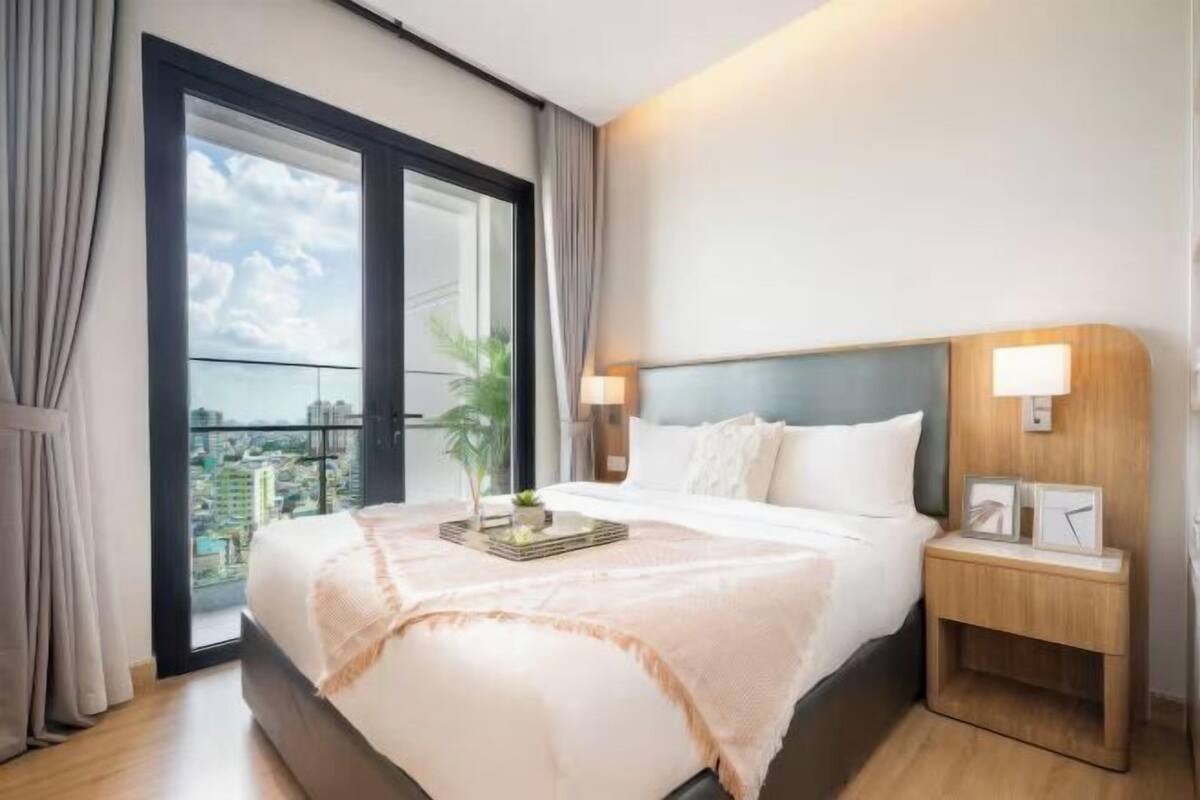 Una habitación de hotel con una cama grande, una mesita de noche con lámpara y vistas de la ciudad a través de un amplio ventanal.