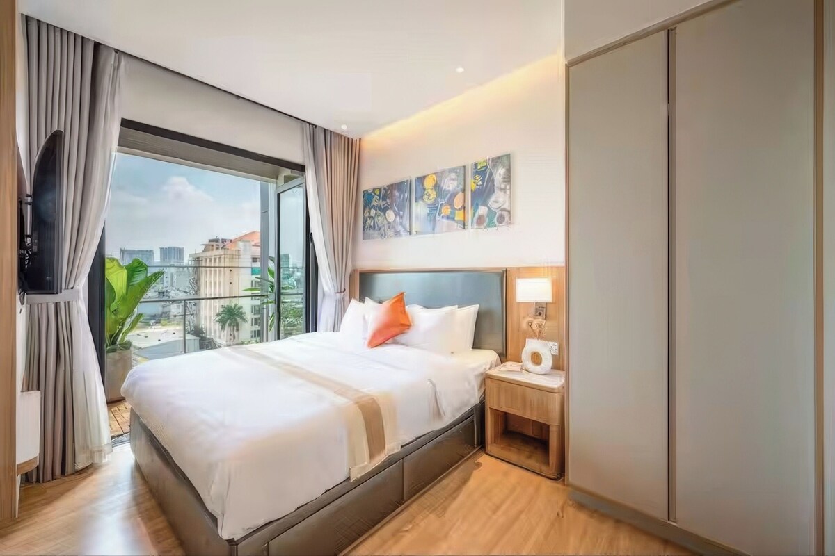A hotel room with a large bed, a TV, a nightstand with a lamp, and a city view.