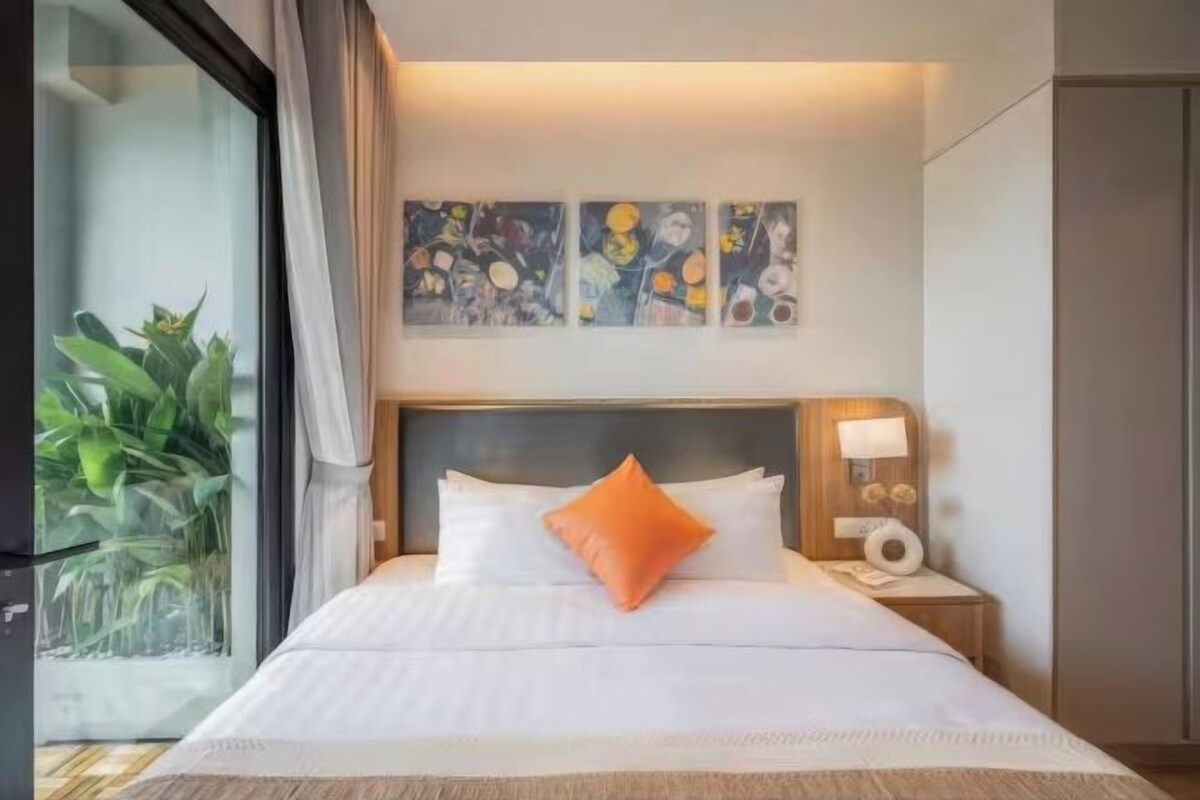 A hotel room with a bed, a nightstand, a lamp, and a window with a view of greenery.