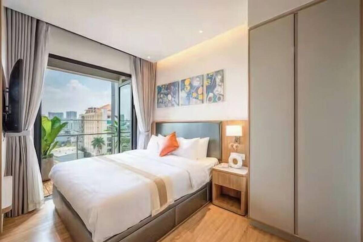 Habitación de hotel con una cama grande, una mesita de noche con lámpara, televisor y ventana con cortinas.