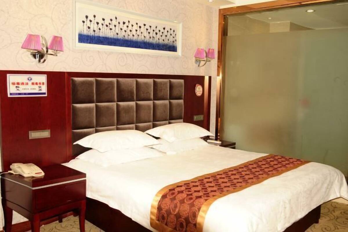 A hotel room with a large bed, a bedside table with a phone, a wall-mounted lamp, and a framed picture above the bed.