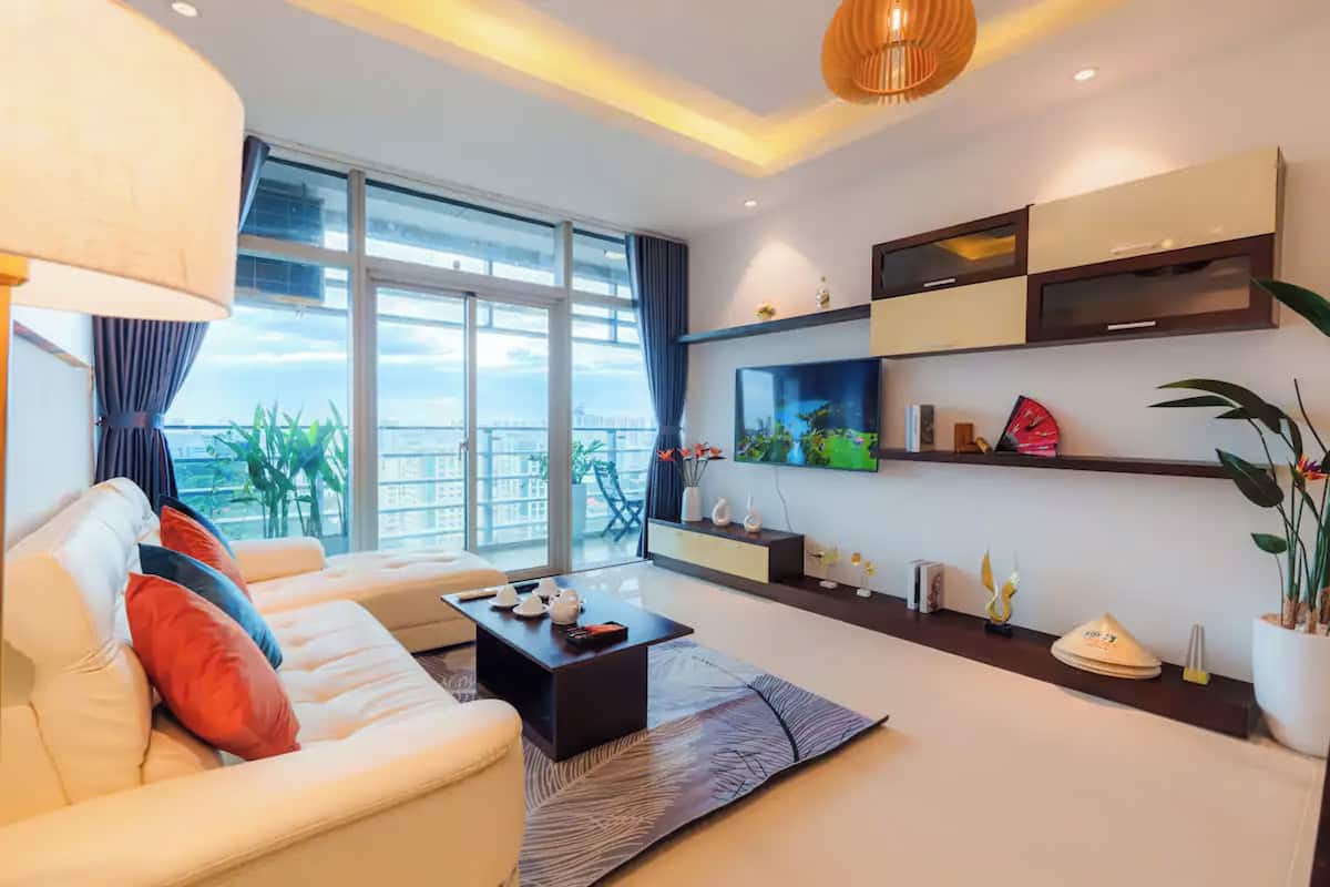 Ein modernes Wohnzimmer mit einer Couch, einem Couchtisch und einem Fernseher. Ein Balkon mit Pflanzen bietet Blick auf die Stadt.