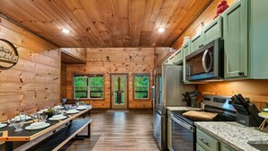A wooden interior with a long dining table set for four, a kitchen with green cabinets, and a view through the door. - 
Smoky Mountain Cabin; 3 King Suites; Hot Tub; Game Room; Sleeps 12 (Sevierville)