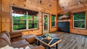 A wooden cabin interior with a brown leather sofa, a coffee table with a decorative centerpiece, and large windows offering a view of the forest. - 
Smoky Mountain Cabin; 3 King Suites; Hot Tub; Game Room; Sleeps 12 (Sevierville)