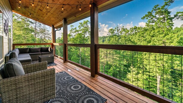A covered outdoor deck with wicker furniture and a view of a forest. - 
Smoky Mountain Cabin; 3 King Suites; Hot Tub; Game Room; Sleeps 12 (Sevierville)