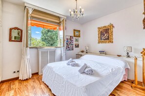 Un dormitorio con una cama grande, una ventana con cortinas, una araña y cuadros enmarcados en la pared.