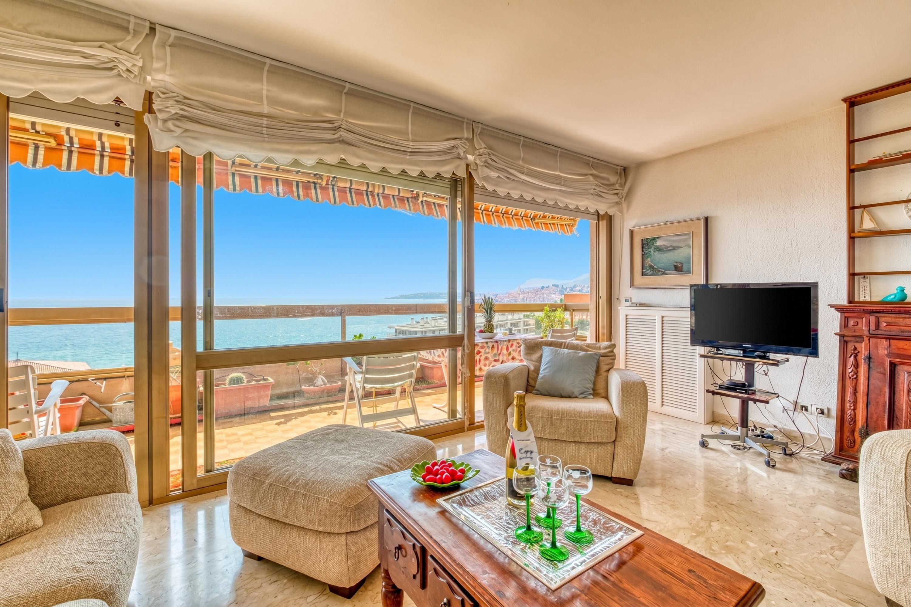 A living room with a view of the sea, a television, and a dining area with a table set for two.