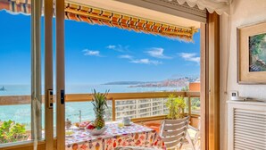 A balcony with a table set for dining, a chair, a potted plant, and a view of the sea and buildings.