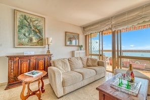 A living room with a beige sofa, a wooden cabinet, a table with a book and a bottle, and a painting on the wall. - La Cuse Baciccia, Menton, France (Menton)