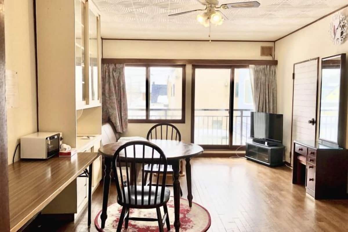 A room with a dining table, chairs, a ceiling fan, a television, and a sliding door to a balcony.