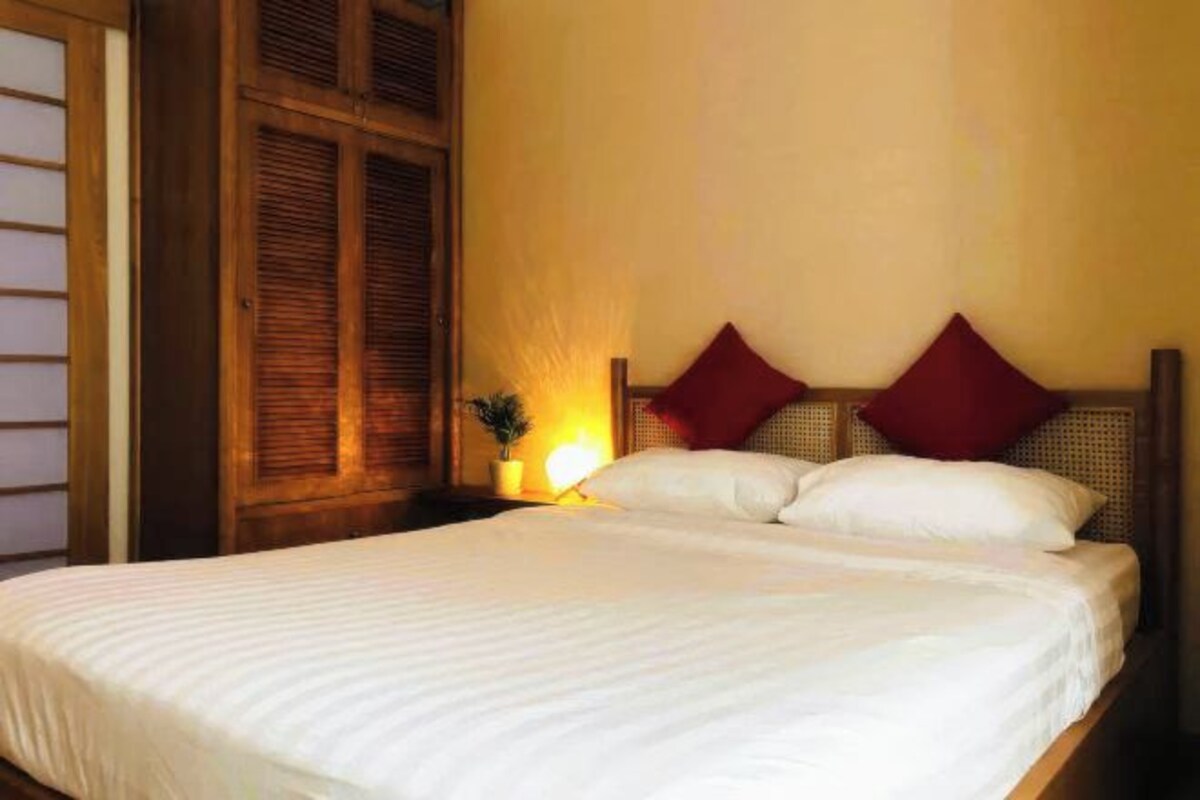 Una cama con ropa blanca, una mesita de noche con lámpara y un cabecero de madera con dos almohadas rojas.