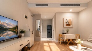 Una sala de estar moderna con una televisión de pantalla plana, piso de madera, un comedor con mesa y sillas, y una cocina con refrigerador.