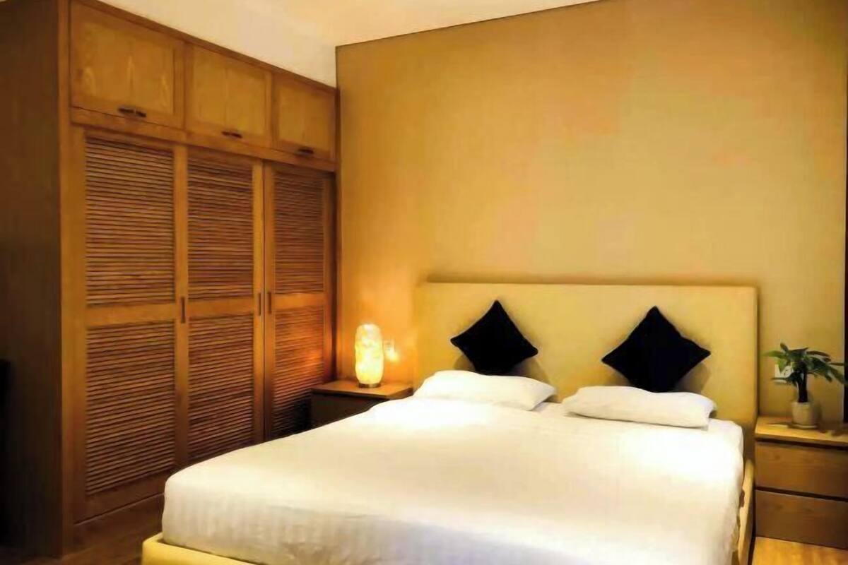 A bedroom with a bed, wooden wardrobe, bedside table with a lamp, and a nightstand with a plant.