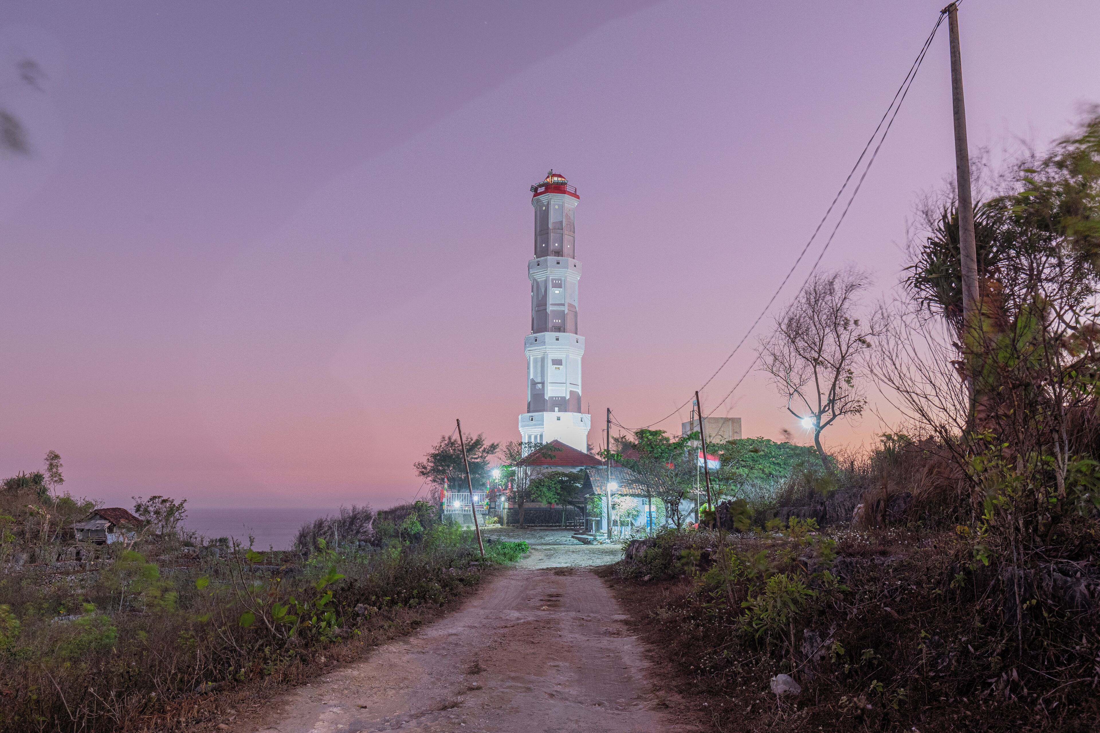 A lighthouse with a clear path leading to it, surrounded by vegetation and a few buildings in the background.
