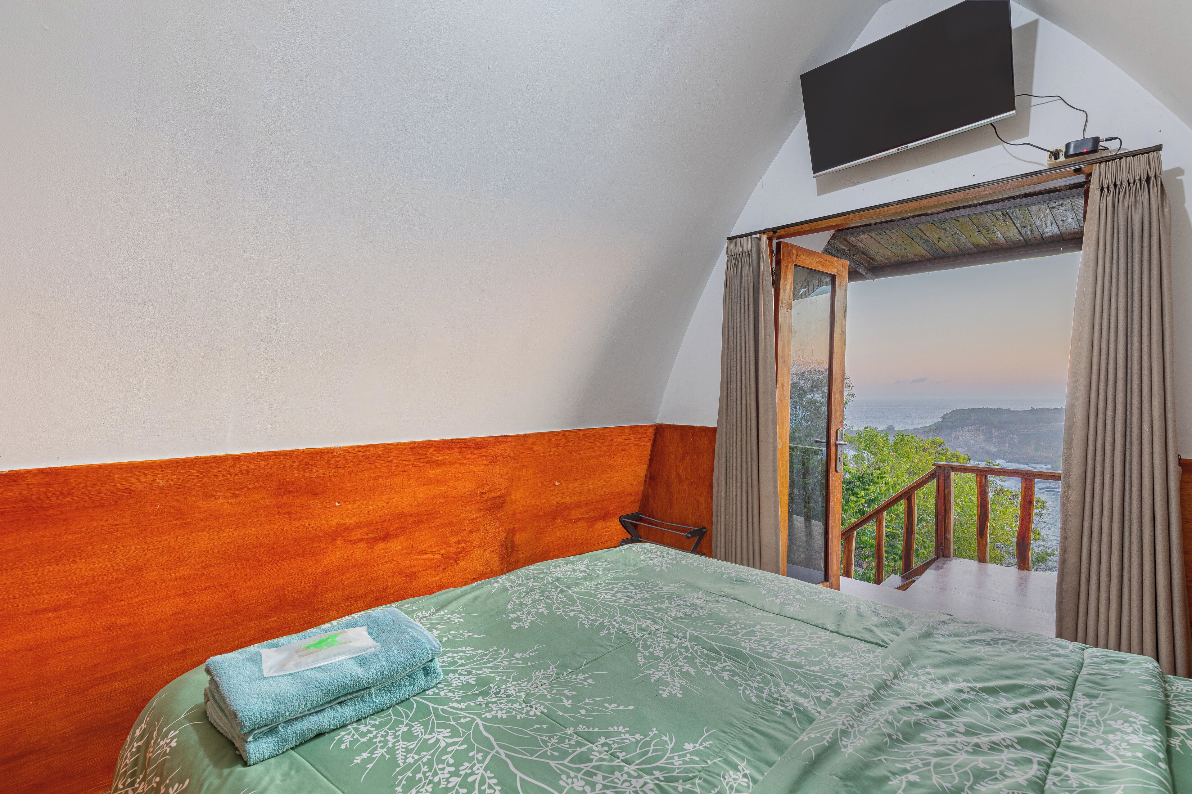 A bedroom with a green floral bedspread, a wooden headboard, a TV mounted on the wall, and a balcony with a view of the sea and cliffs.