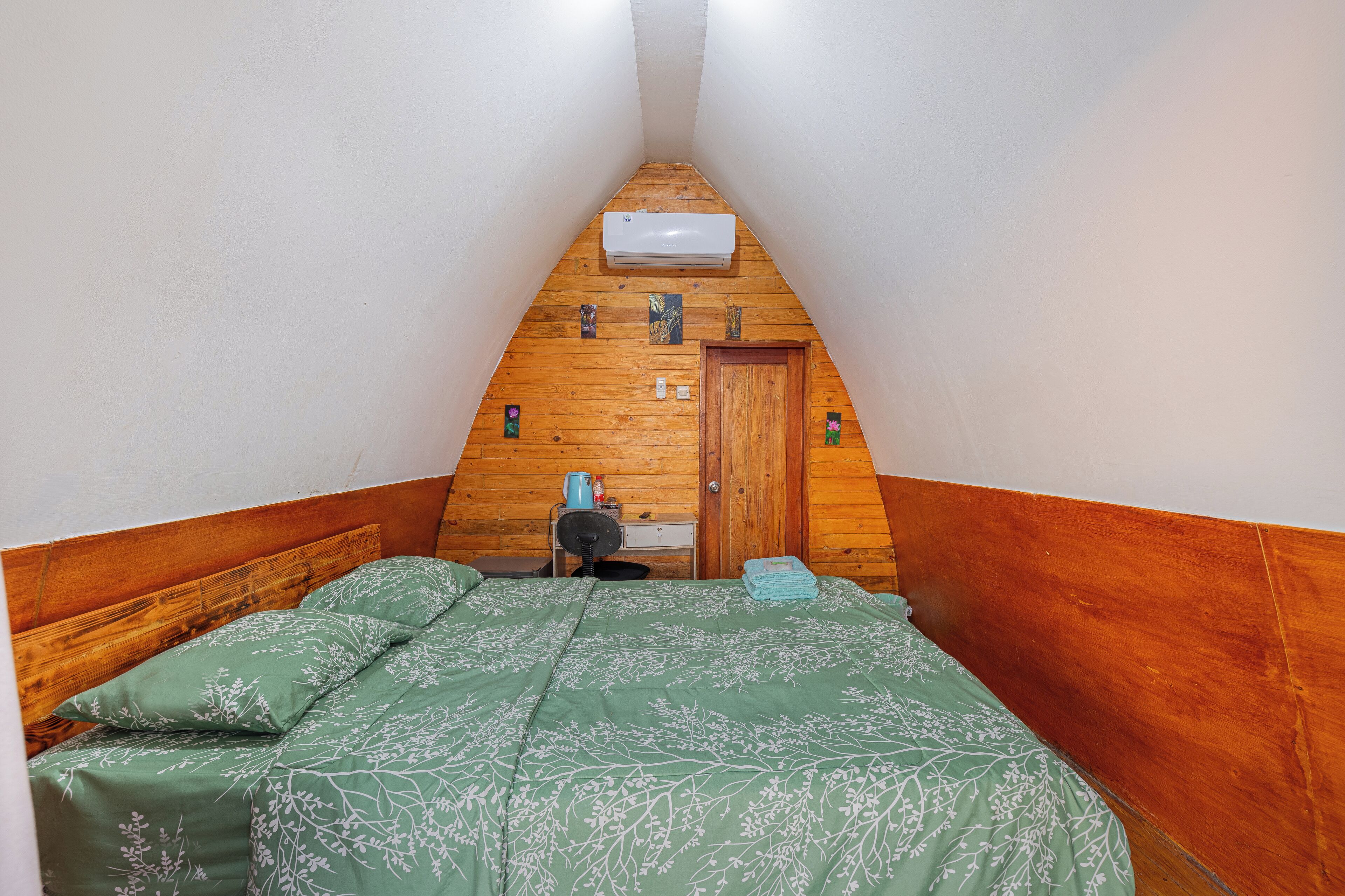 Um quarto pequeno e triangular com teto inclinado de madeira, uma cama com roupa de cama floral verde, uma escrivaninha de madeira com uma cadeira e uma pequena janela.