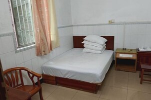 Una cama individual con cabecera de madera, una silla de madera, una mesita de noche pequeña y una ventana con cortina.