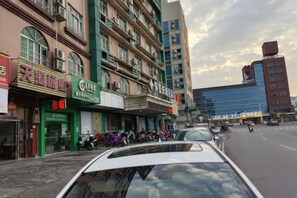 Vista de la calle con varias tiendas, un coche estacionado y un edificio de fachada azul.
