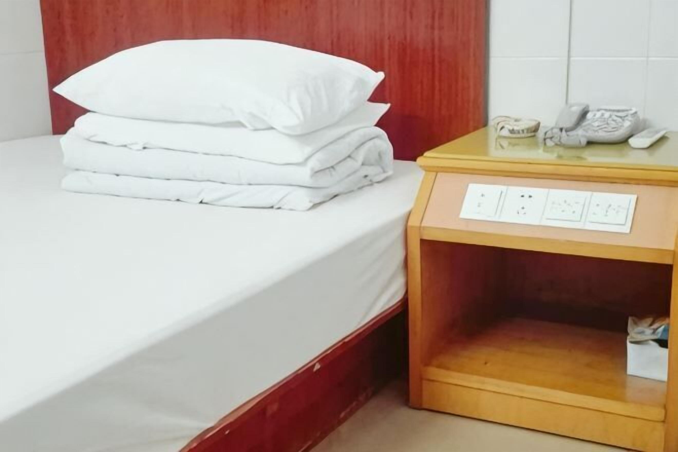 A neatly made bed with white linens and pillows, a wooden nightstand with a telephone and a power outlet, and a tiled wall in the background.