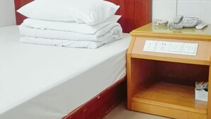 A neatly made bed with white linens and pillows, a wooden nightstand with a telephone and a power outlet, and a tiled wall in the background. - Huaiji Tianyuan Travel Industry (Huaiji)