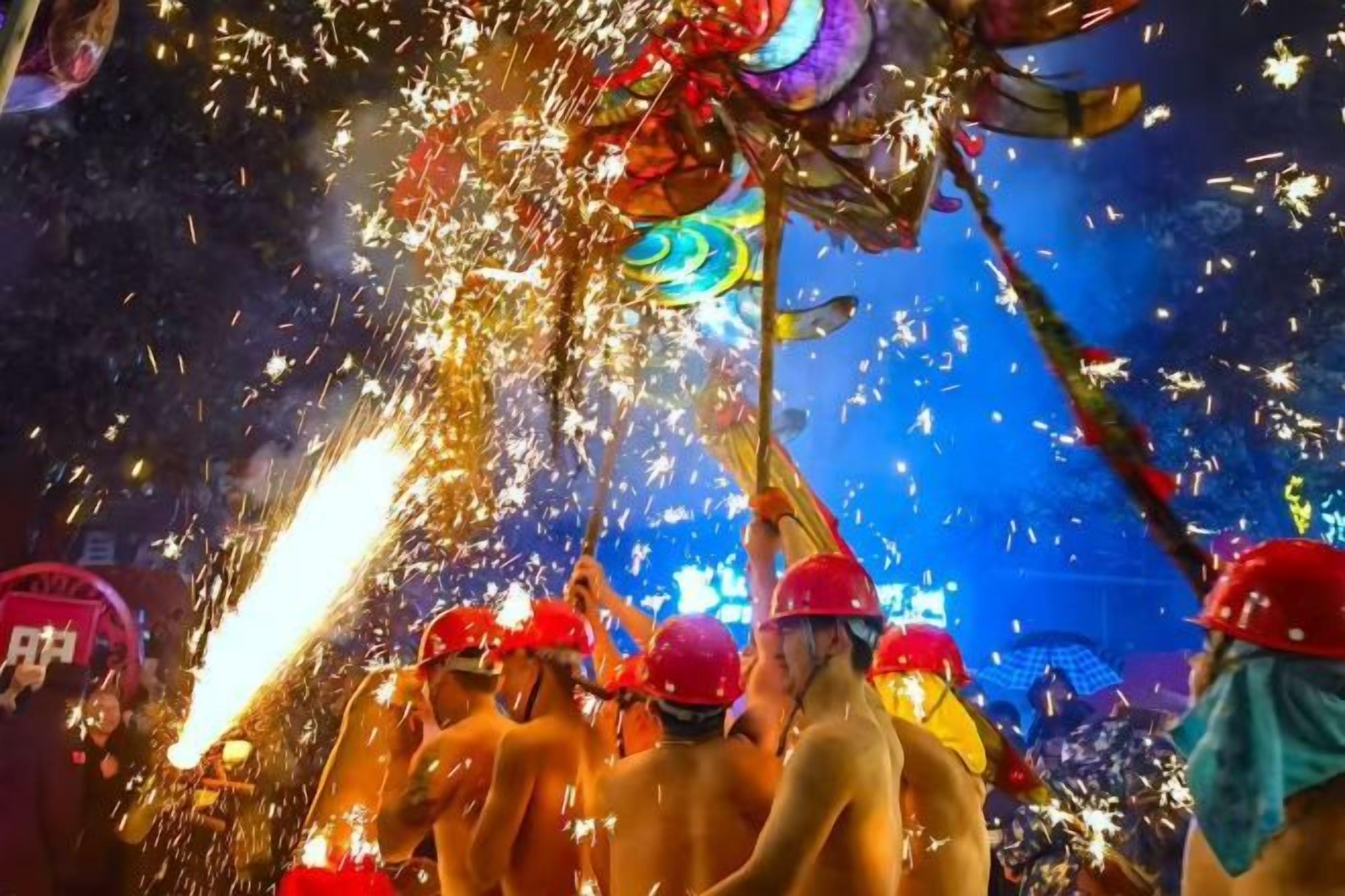 Un grupo de personas con cascos rojos y ropa naranja participan en una actividad que genera chispas junto a una estructura grande.