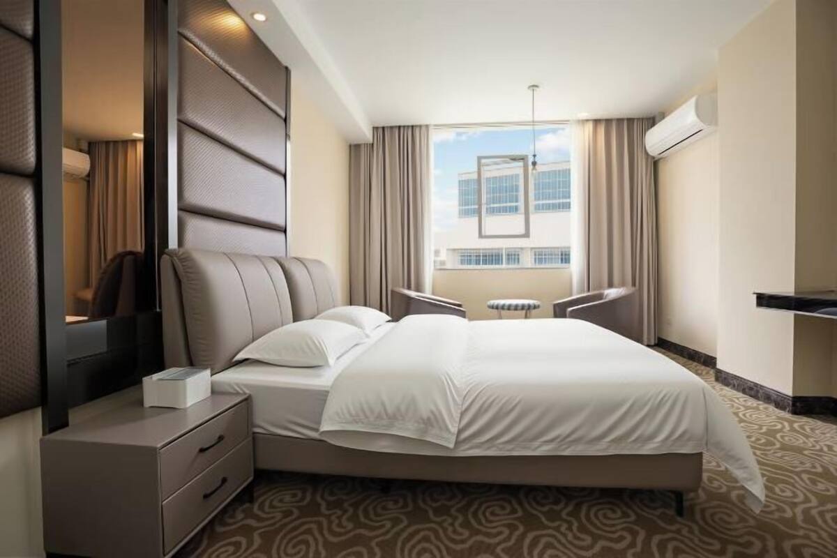 Ein Hotelzimmer mit einem großen Bett, einem Nachttisch, einem Sessel, einem Fenster mit Vorhängen und einer Klimaanlage.