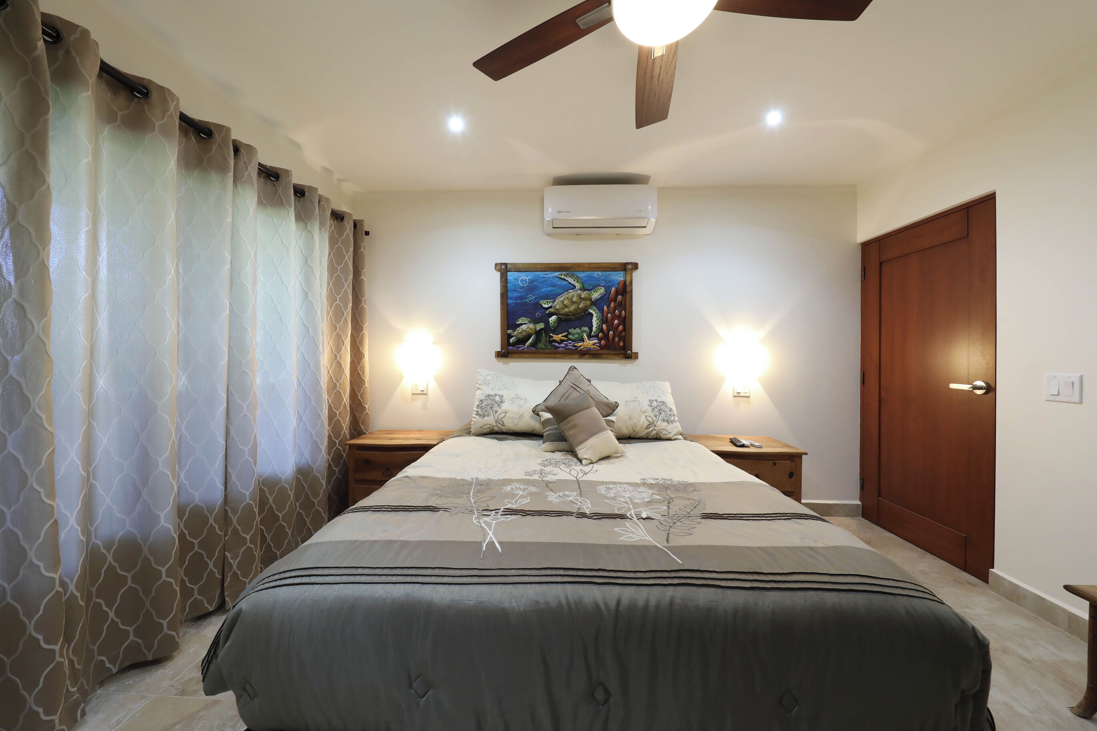 Un dormitorio con cama, mesitas de noche, ventilador de techo, aire acondicionado y un cuadro en la pared.