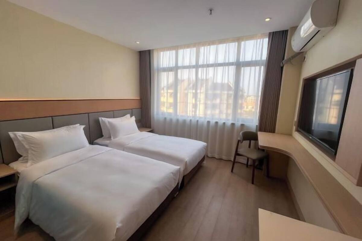 Een hotelkamer met twee bedden, een bureau, een stoel, een groot raam met gordijnen en een airconditioning.