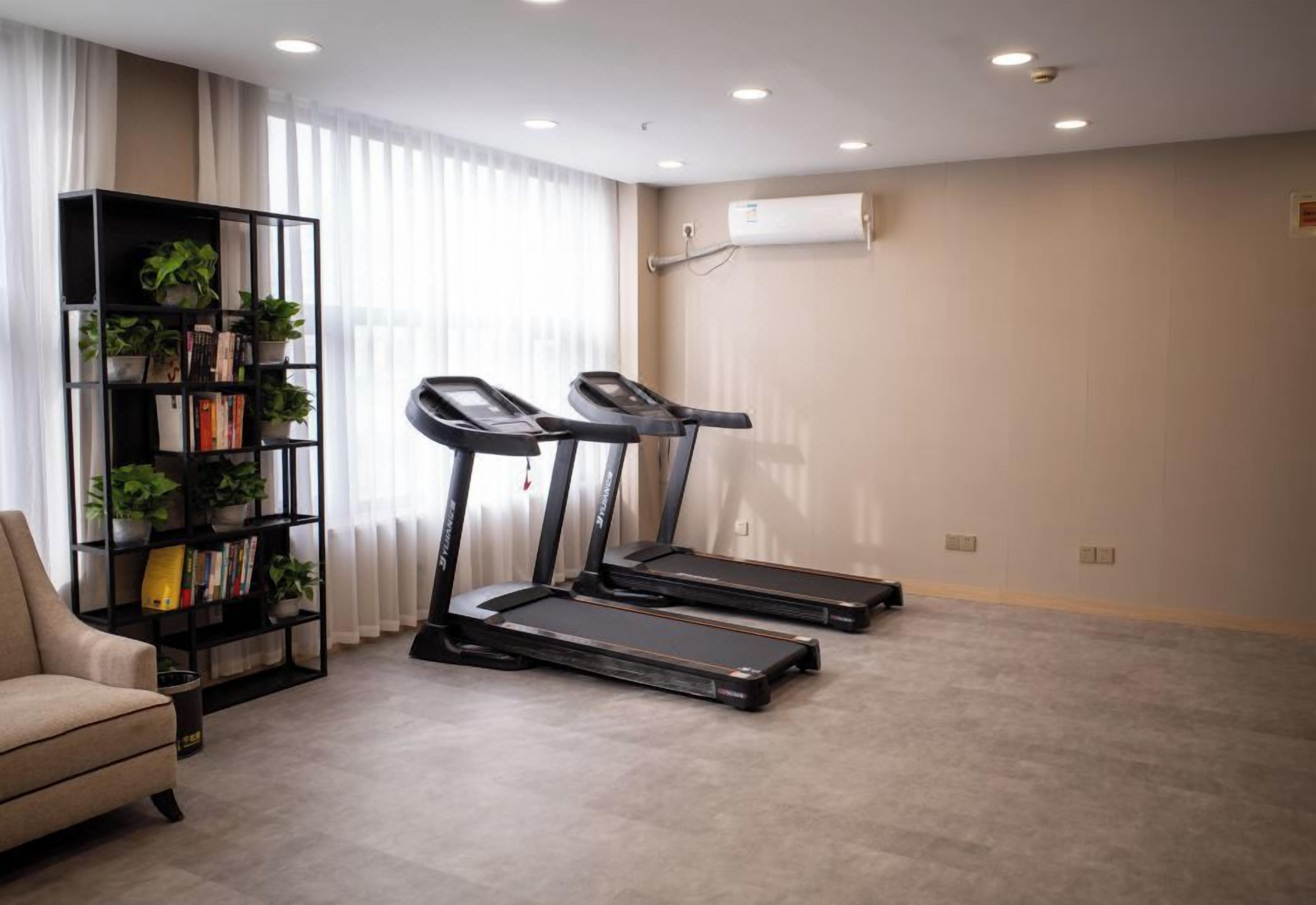 A room with a treadmill, a bookshelf with plants, and a beige armchair.