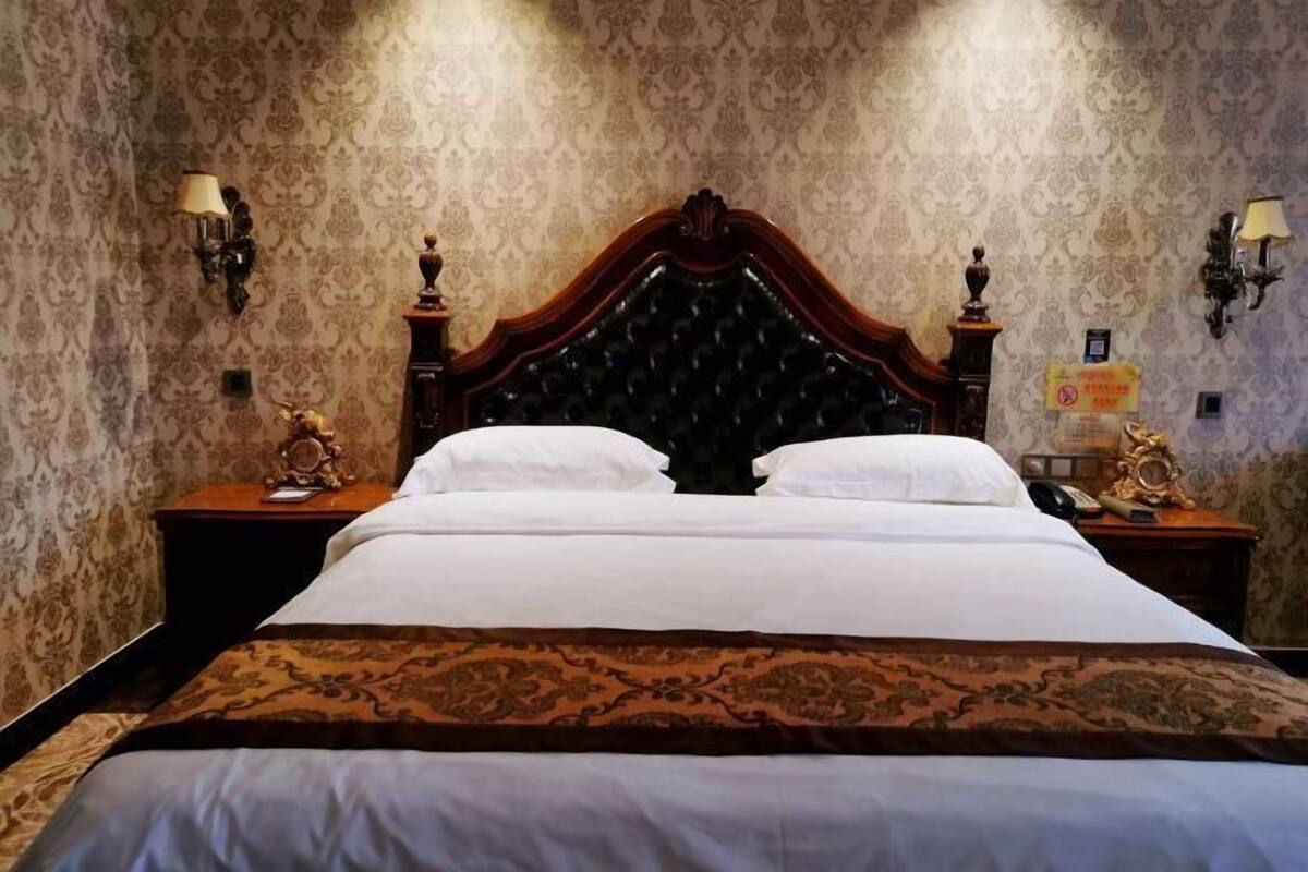 A hotel room with a large bed, ornate headboard, and patterned wallpaper. There are two bedside tables with lamps and decorative items.