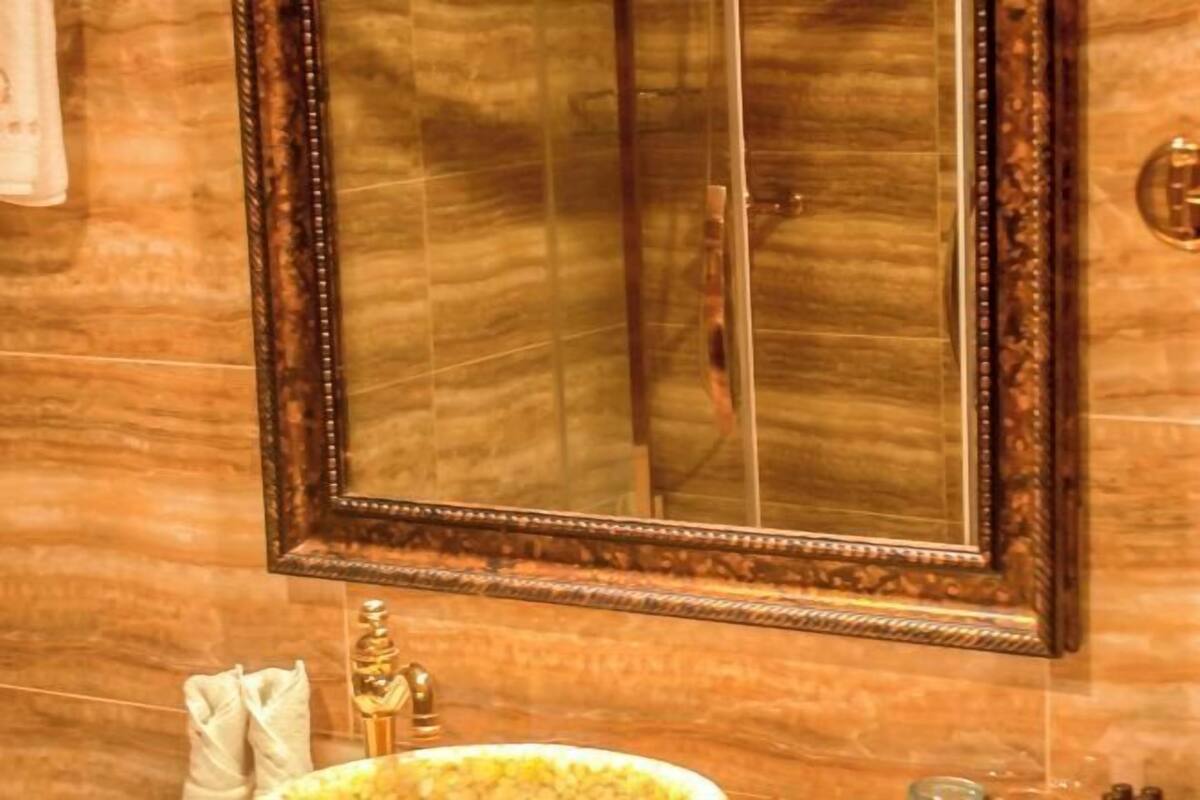 A bathroom with a marble wall, a large mirror with a wooden frame, a gold faucet, and a towel hanging on the wall.